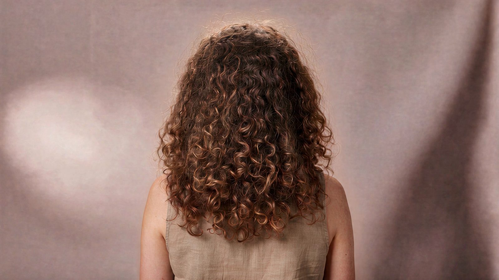 The back of a woman's head, long natural curls at rest against a pale blush studio backdrop